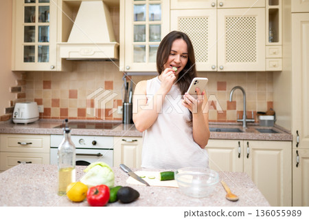 A young woman in a cozy kitchen eats a cucumber slice while checking her phone. Fresh vegetables on the countertop. Healthy lifestyle, home cooking, and balanced nutrition. 136055989