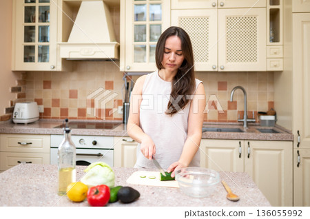 Smiling woman cutting cucumber for fresh vegetable salad in cozy kitchen. Healthy lifestyle, home cooking, diet and proper nutrition 136055992