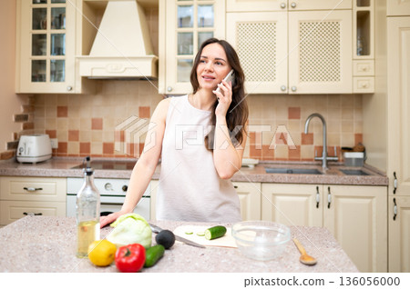 young woman in bright kitchen talking on phone and smiling, planning to cook salad. Fresh vegetables and board are on table young woman in bright kitchen talking on phone and smiling, planning to cook salad. Fresh vegetables and board are on table 136056000