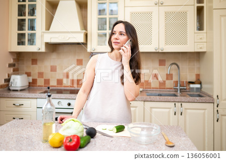 young woman in bright kitchen talking on phone and smiling, planning to cook salad. Fresh vegetables and board are on table young woman in bright kitchen talking on phone and smiling, planning to cook salad. Fresh vegetables and board are on table 136056001