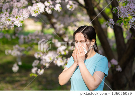 Woman sneezing into tissue in blooming garden, seasonal allergy symptoms during springtime Woman sneezing into tissue in blooming garden, seasonal allergy symptoms during springtime 136056089