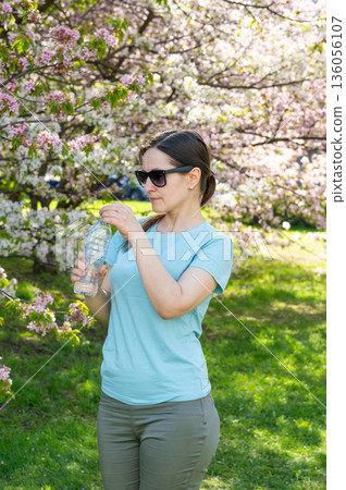 Young woman wearing sunglasses drinking water from bottle outdoors in blooming spring park Young woman wearing sunglasses drinking water from bottle outdoors in blooming spring park 136056107