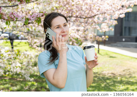 Young woman talking on smartphone and drinking hot drink in blooming spring park, coffee break Young woman talking on smartphone and drinking hot drink in blooming spring park, coffee break 136056164