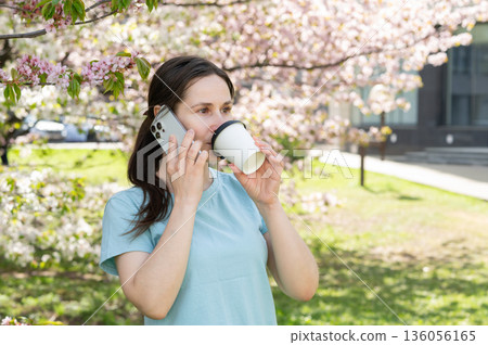 Relaxed young woman enjoys hot beverage and phone call while sitting in blooming spring park Relaxed young woman enjoys hot beverage and phone call while sitting in blooming spring park 136056165