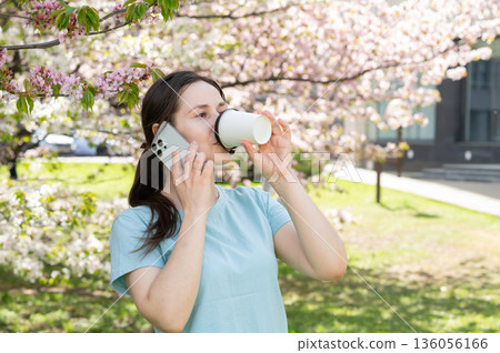 Young woman talking on smartphone and drinking hot drink in blooming spring park, coffee break Young woman talking on smartphone and drinking hot drink in blooming spring park, coffee break 136056166
