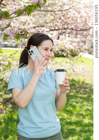 Relaxed young woman enjoys hot beverage and phone call while sitting in blooming spring park 136056195