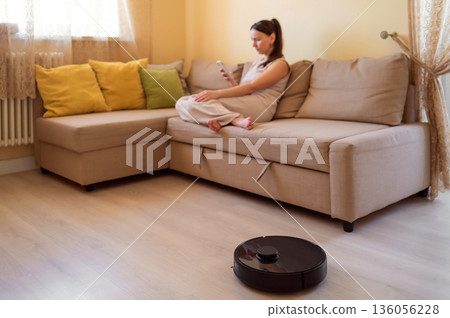A robot vacuum cleaner washes the floor while a young woman uses her smartphone while sitting on a cozy beige sofa. Smart home, automation and modern technologies in everyday life A robot vacuum cleaner washes the floor while a young woman uses her smartphone while sitting on a cozy beige sofa. Smart home, automation and modern technologies in everyday life 136056228