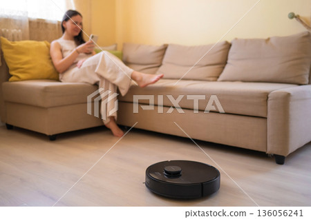 A robot vacuum cleaner washes the floor while a young woman uses her smartphone while sitting on a cozy beige sofa. Smart home, automation and modern technologies in everyday life A robot vacuum cleaner washes the floor while a young woman uses her smartphone while sitting on a cozy beige sofa. Smart home, automation and modern technologies in everyday life 136056241