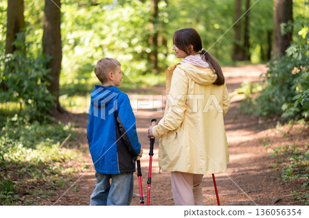 Rear view of mother and 11-year-old son hiking with Nordic trekking poles through a sunny forest trail 136056354