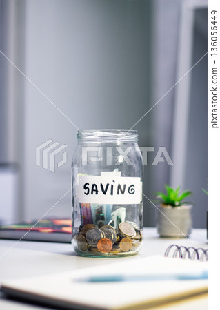 Hand dropping coins into glass savings jar on table. Close up of fingers inserting money into transparent container, representing simple financial planning and goal achievement in everyday life 136056449