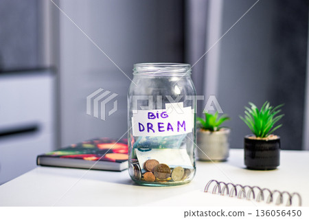 Person saving money in glass jar for future dreams and dropping coins into transparent savings jar. Symbolizes goal setting, financial planning 136056450