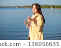 Woman in a yellow raincoat holding dried flowers by the lake, enjoying the warm rays of the sun 136056635
