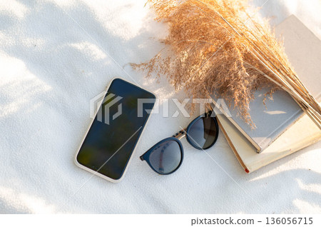Still life with smartphone, books, dried flowers, and sunglasses on a white blanket in warm natural light, evoking a peaceful weekend vibe 136056715