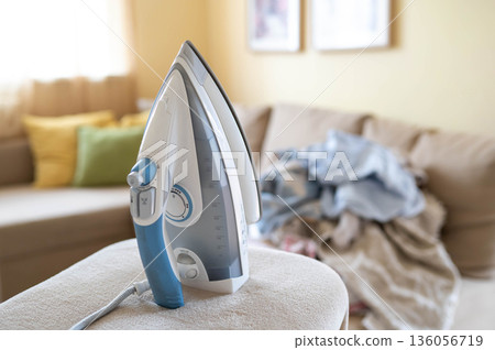 Unplugged iron with power cord and plug on ironing board, blurred background with wrinkled laundry on sofa Unplugged iron with power cord and plug on ironing board, blurred background with wrinkled laundry on sofa 136056719