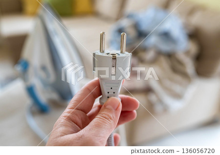 A plug with a power cord from a disconnected iron in a woman's hand against the background of an ironing board with an iron and a pile of crumpled laundry A plug with a power cord from a disconnected iron in a woman's hand against the background of an ironing board with an iron and a pile of crumpled laundry 136056720