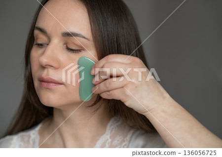 Young woman using a green gua sha scraper for facial massage, improving skin circulation and relaxation. Home skincare and self-care routine for beauty and wellness. 136056725