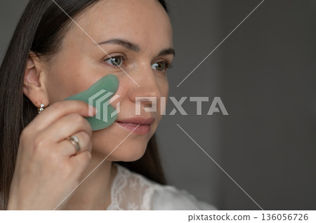 Young woman using a green gua sha scraper for facial massage, improving skin circulation and relaxation. Home skincare and self-care routine for beauty and wellness. 136056726