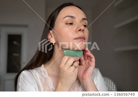 Young woman in silk robe doing facial massage with gua sha scraper while looking in mirror. Home skin care and aesthetic procedures 136056749