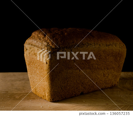 Loaf of black rye bread, in the shape of a brick on a wooden board on a black background Loaf of black rye bread, in the shape of a brick on a wooden board on a black background 136057235