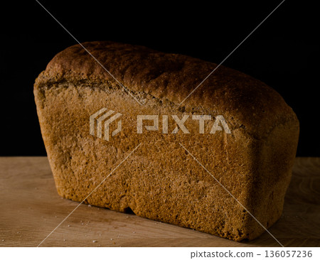 Loaf of black rye bread, in the shape of a brick on a wooden board on a black background Loaf of black rye bread, in the shape of a brick on a wooden board on a black background 136057236