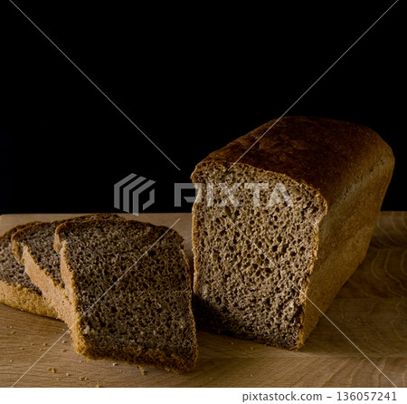 Loaf of black rye bread, cut into pieces, in the shape of a brick on a wooden board on a black background 136057241