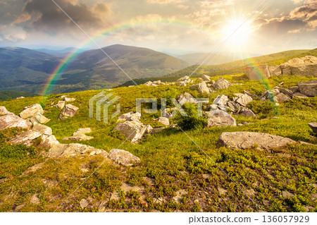 mountain landscape in summer with rocks at sunset. green grass under blue sky in dramatic evening light. alpine meadow and rolling hills with boulders. timeless nostalgia book cover with rainbow 136057929