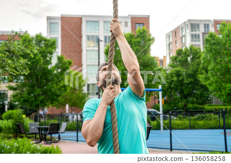 Strong man climbing on the rope during training in sport gym. Mature man athlete climbing a rope. Man do workout exercise. Fitness rope climb exercise in fitness gym workout outdoor 136058258