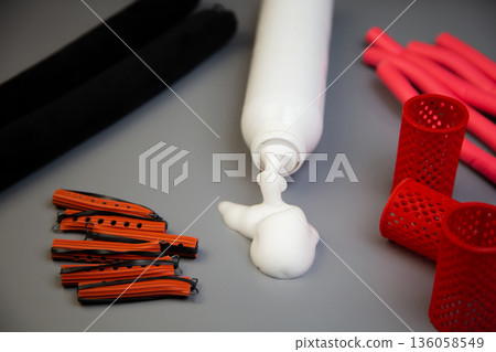 A close-up on a gray background shows a bottle of foam for long-term hair styling, white foam poured out of it, next to it there are curlers of different shapes and sizes. The concept of styling A close-up on a gray background shows a bottle of foam for long-term hair styling, white foam poured out of it, next to it there are curlers of different shapes and sizes. The concept of styling 136058549