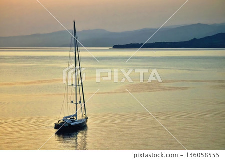 A single yacht sailing on calm waters in the early morning sunrise with distant mountains and hills in the distance A single yacht sailing on calm waters in the early morning sunrise with distant mountains and hills in the distance 136058555