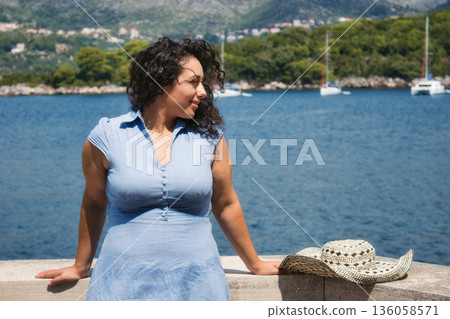A beautiful white Caucasian woman with curly black hair wearing a blue dress, on vacation at the beach, with water and yachts visible in the background 136058571