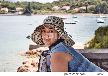 A beautiful white Caucasian woman wearing a bit floppy summer straw hat, looking directly at camera, against a backdrop of boats in the water on a Mediterranean island 136058575