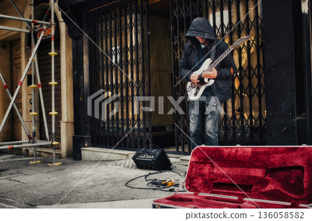 An unrecognizable hooded busker in a pedestrian street in a city center playing an electric blues guitar with an amplifier and effects pedals An unrecognizable hooded busker in a pedestrian street in a city center playing an electric blues guitar with an amplifier and effects pedals 136058582