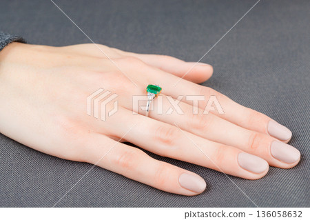 Beautiful female hand with gold ring with diamonds and emerald on gray background Beautiful female hand with gold ring with diamonds and emerald on gray background 136058632