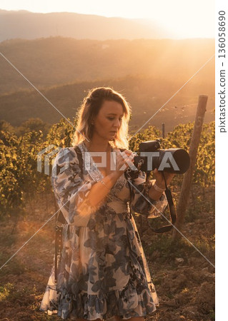 Woman in floral dress using camera in a lush vineyard during golden hour. Her focus on the camera suggests engagement with her surroundings. Peaceful mountain background completes the scene. Woman in floral dress using camera in a lush vineyard during golden hour. Her focus on the camera suggests engagement with her surroundings. Peaceful mountain background completes the scene. 136058690