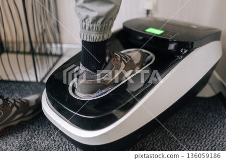 Close-up view of unrecognizable person wearing sneakers using automatic shoe cover dispenser, which wraps protective plastic film over footwear for hygiene and cleanliness in sterile environment. Close-up view of unrecognizable person wearing sneakers using automatic shoe cover dispenser, which wraps protective plastic film over footwear for hygiene and cleanliness in sterile environment. 136059186