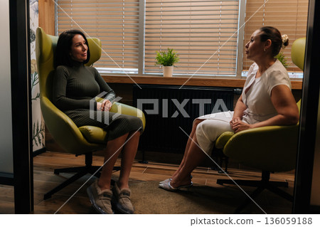 Side view of female client having confidential one-on-one session with professional psychotherapist, discussing mental health concerns and receiving counseling in dark private office setting. Side view of female client having confidential one-on-one session with professional psychotherapist, discussing mental health concerns and receiving counseling in dark private office setting. 136059188
