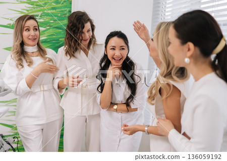 Cheerful five female healthcare professionals in white uniforms gathered together in modern clinic office, exchanging smiles as they discuss and share their expertise during medical conference. 136059192