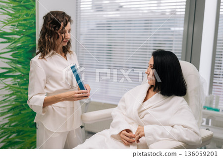 Aesthetician doctor showing product to client sitting in white robe, discussing personalized skincare solutions during professional consultation for health and beauty in contemporary clinic. 136059201