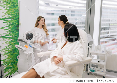 Skilled cosmetic professional doctor explaining advanced beauty treatment technology to client during consultation in modern aesthetic clinic, with another professional assisting. Skilled cosmetic professional doctor explaining advanced beauty treatment technology to client during consultation in modern aesthetic clinic, with another professional assisting. 136059209
