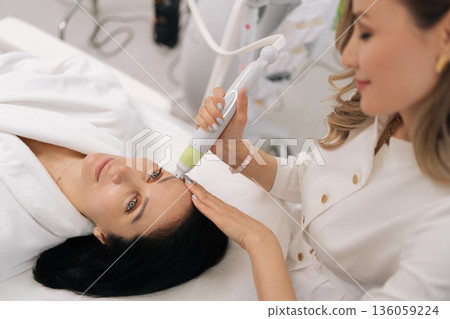 Closeup top view of woman patient receiving professional facial treatment in beauty salon, undergoing hardware mesotherapy and microneedling to rejuvenate skin and achieve anti-aging effect. 136059224