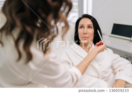Back view of experienced beauty technician consulting with woman seated comfortably in chair, using pencil to outline features for upcoming cosmetic enhancement at contemporary beauty center. 136059233