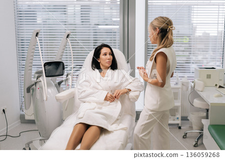 Cosmetologist consulting female client about skin rejuvenation treatment using advanced ultrasound device in modern dermatological clinic, discussing options for beauty care. Cosmetologist consulting female client about skin rejuvenation treatment using advanced ultrasound device in modern dermatological clinic, discussing options for beauty care. 136059268