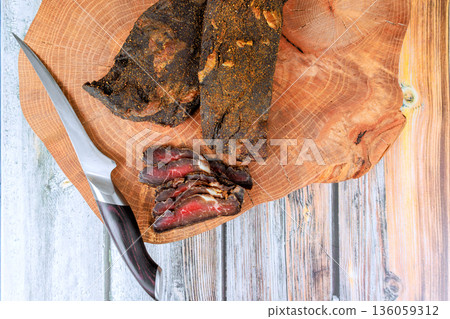 Cured beef meat is sliced arranged on wooden board, knife rests next to meat in kitchen. Cured beef meat is sliced arranged on wooden board, knife rests next to meat in kitchen. 136059312