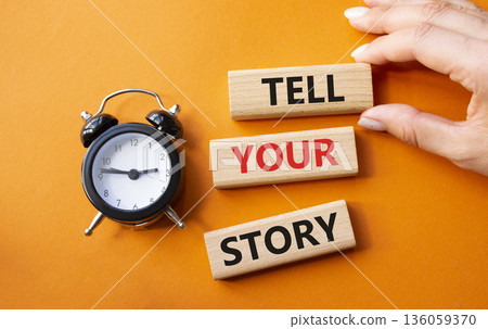 Tell your story symbol. Wooden blocks with words Tell your story. Businessman hand. Beautiful orange background with alarm clock. Business and Tell your story concept. Copy space. 136059370