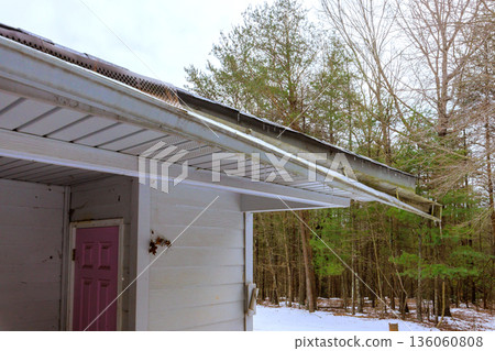 Snow rests on roof broken gutter while trees stand tall in winter morning. 136060808
