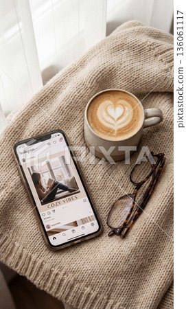 Smartphone showing social media app, latte with heart art and glasses on beige knitwear. Cozy warm atmosphere for lifestyle blogging, relaxation themes and social media marketing. Smartphone showing social media app, latte with heart art and glasses on beige knitwear. Cozy warm atmosphere for lifestyle blogging, relaxation themes and social media marketing. 136061117