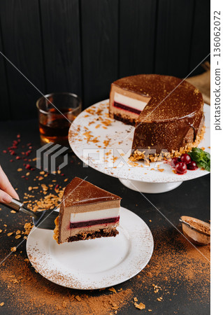 Layered chocolate mousse cake slice with raspberry jelly on spatula, dark background Layered chocolate mousse cake slice with raspberry jelly on spatula, dark background 136062072