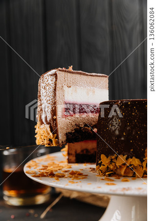 Layered chocolate mousse cake slice with raspberry jelly on spatula, dark background Layered chocolate mousse cake slice with raspberry jelly on spatula, dark background 136062084