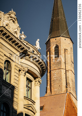 Historic Church Tower And Ornate Facade In Munich: Tall Stone Spire Rising Beside Decorative Classical Architecture Under Clear Blue Sky 136062356