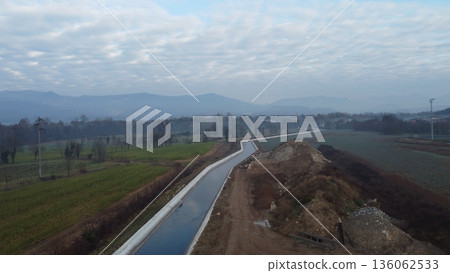 A professional drone shot of a modern concrete water canal crossing agricultural fields in Bedizzole, Brescia province. The background features the Prealpine mountains of Lombardy A professional drone shot of a modern concrete water canal crossing agricultural fields in Bedizzole, Brescia province. The background features the Prealpine mountains of Lombardy 136062533